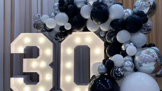 Chiffre lumineux géant 30 en carton mousse blanc éclairé devant une arche de ballons noirs, blancs et argentés