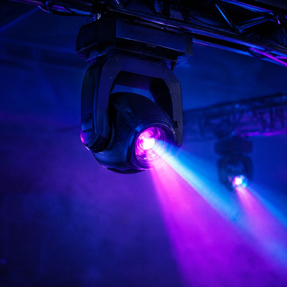 Lyre LED suspendue à une structure noire projetant des faisceaux cyan et magenta dans la brume d'une salle de soirée