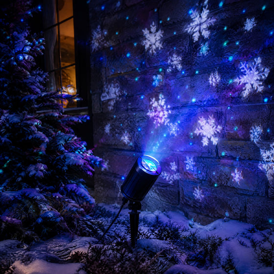 Projecteur de Noël extérieur planté dans un jardin enneigé, flocons et points lumineux projetés sur la façade d'une maison