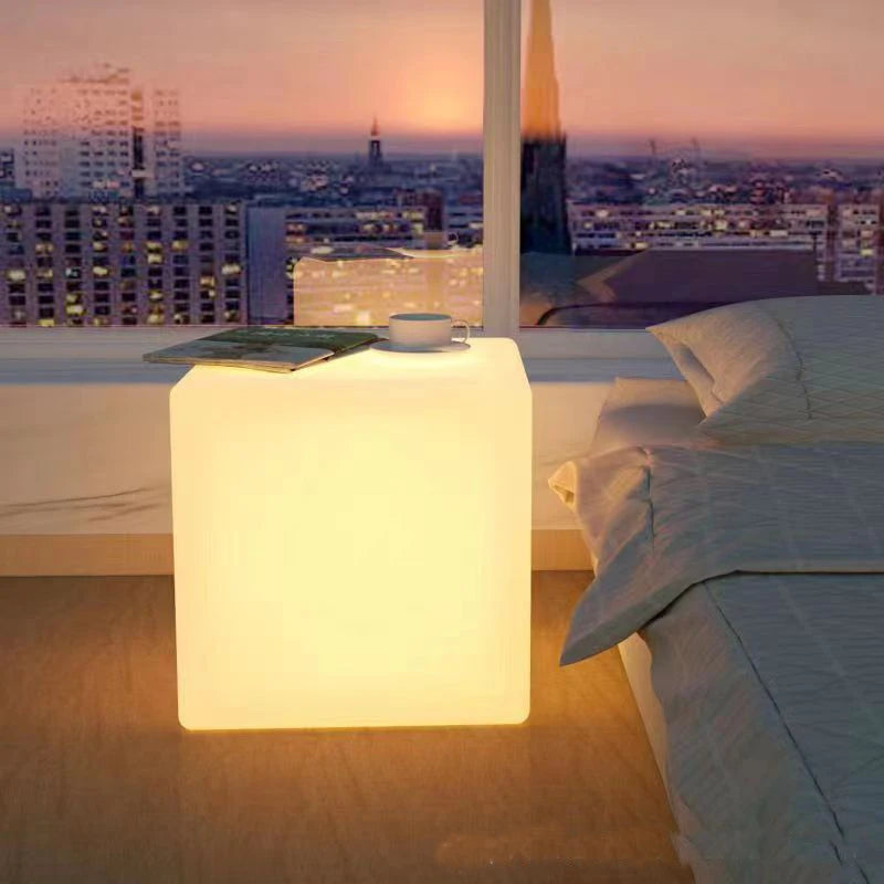 Cube lumineux LED – cube blanc chaud au pied d'un lit devant une fenêtre, magazine et tasse posés dessus
